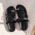 A pair of black leather slide sandals with a small silver metal accent on the front strap, displayed on a white fabric surface.