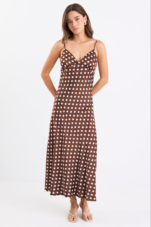 Woman wearing a chocolate brown satin midi dress with white polka dots, adjustable spaghetti straps, and a bias-cut silhouette, standing against a white background.