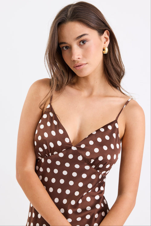 Woman wearing a chocolate brown satin midi dress with white polka dots, adjustable spaghetti straps, and a bias-cut silhouette, standing against a white background.
