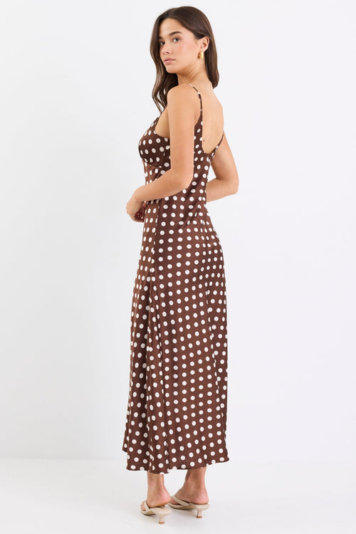 Woman wearing a chocolate brown satin midi dress with white polka dots, adjustable spaghetti straps, and a bias-cut silhouette, standing against a white background.