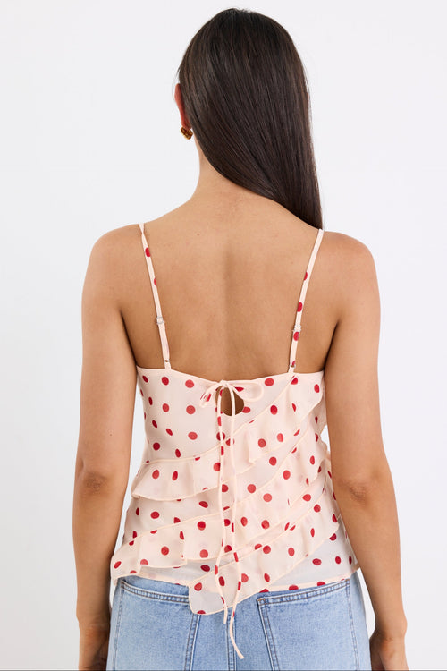 Woman wearing a cream cami top with cherry-red polka dots, ruffled front detailing, and thin adjustable straps, paired with light blue jeans against a white background.