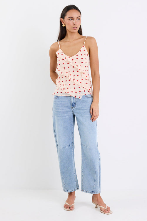 Woman wearing a cream cami top with cherry-red polka dots, ruffled front detailing, and thin adjustable straps, paired with light blue jeans against a white background.