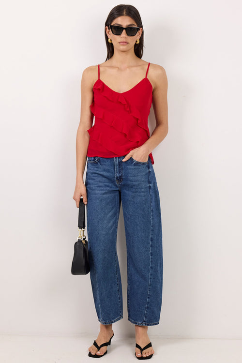 A model stands against a plain white studio background wearing a bright red ruffled spaghetti-strap camisole top, blue wide-leg cropped jeans, black sandals, and holding a small black handbag.