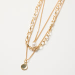 Gold triple-layer necklace with mixed chains, a pearl accent, and a round gold disc charm, styled flat on a white background
