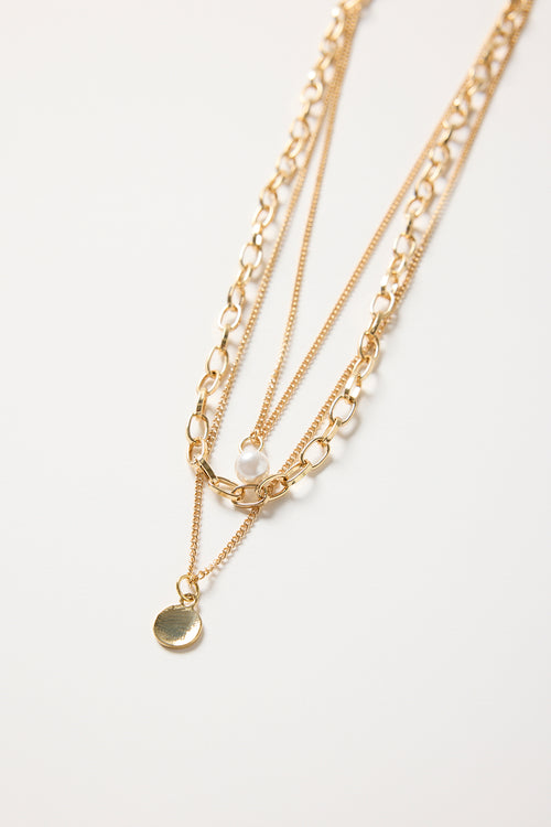 Gold triple-layer necklace with mixed chains, a pearl accent, and a round gold disc charm, styled flat on a white background