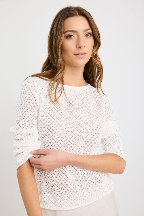 Woman wearing an ivory crochet knit jumper with boat neckline and long sleeves, styled with a natural skirt.