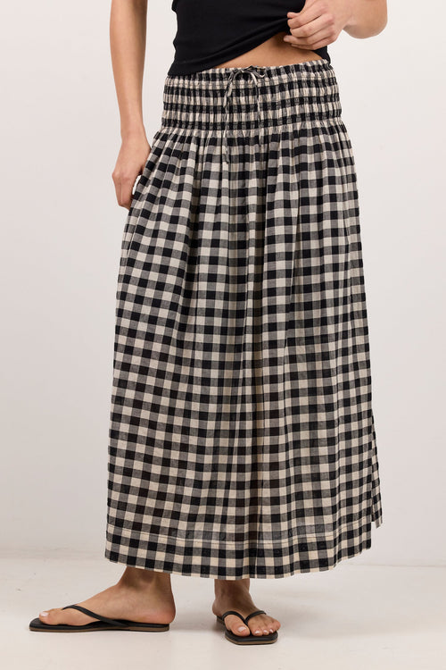 A person wearing a black and beige gingham pleated maxi skirt with a drawstring waist, styled with a black top and black flip-flop sandals against a plain off-white studio background.