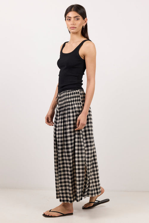 A woman modeling a black sleeveless tank top and black-and-white gingham checkered wide-leg pants, paired with black flip-flop sandals, against a plain white studio background.