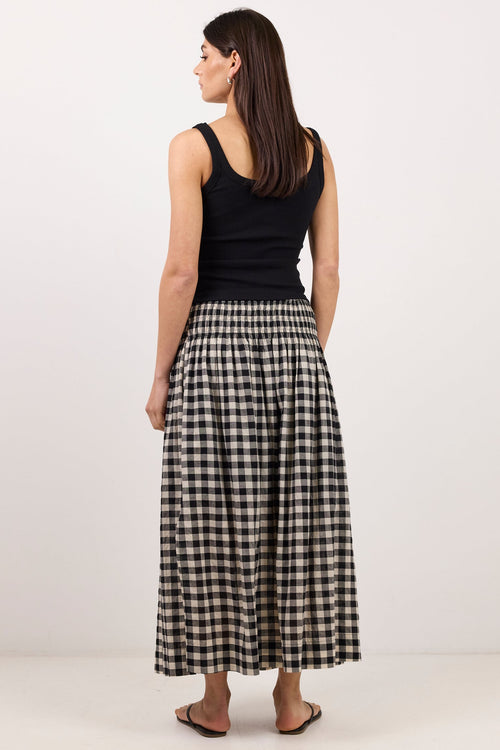 A woman stands with her back to the camera wearing a black sleeveless top and a black-and-white gingham pleated maxi skirt, set against a plain off-white studio background.