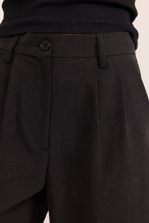Close-up of black tailored trousers showing the waistband, button closure, belt loops, and front pleats, worn with a black ribbed top against a light neutral background.