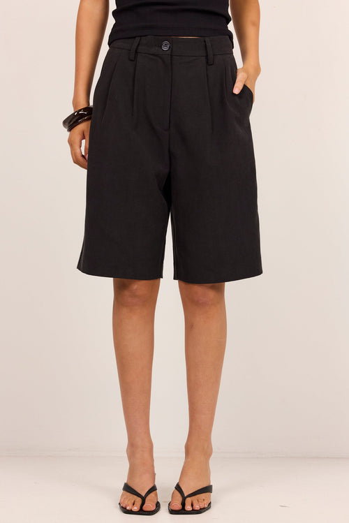 A model from the waist down wearing black pleated knee-length shorts with side pockets, paired with black sandals and a black top, standing against a plain off-white studio background.