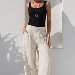 A woman wearing a black sleeveless tank top and cream wide-leg linen trousers stands in a sunlit white-walled courtyard next to a large terracotta planter with a palm plant.