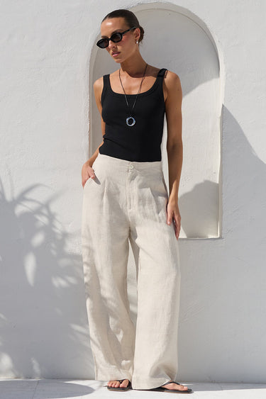 A woman wearing a black sleeveless tank top and cream wide-leg linen trousers stands in a sunlit white-walled courtyard next to a large terracotta planter with a palm plant.