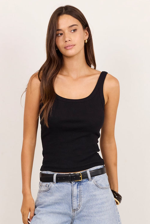A woman modeling a black ribbed tank top, light-wash cropped denim culottes, a black belt, and black slide sandals against a plain off-white studio background.