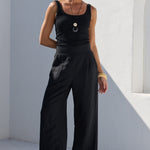 A woman wearing a black sleeveless top and matching wide-leg pants stands in a minimalist white architectural setting with strong sunlight and shadows.