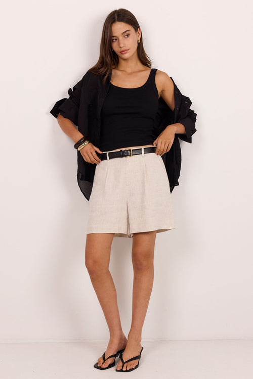 A woman modeling a black cold-shoulder top and open black jacket over a black camisole, paired with light beige shorts and black sandals, standing against a plain white studio background.