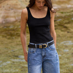 A woman standing outdoors on a stone ledge next to a shallow stream, wearing a black sleeveless top, blue denim cutoff shorts, a black belt with a gold buckle, and black flip-flops.