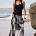 A woman stands barefoot on a rocky beach wearing a black sleeveless tank top and a long black-and-white gingham maxi skirt, with ocean water and large rocks in the background.