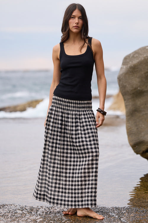 A woman stands barefoot on a rocky beach wearing a black sleeveless tank top and a long black-and-white gingham maxi skirt, with ocean water and large rocks in the background.