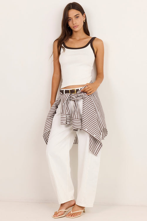A woman stands against a plain off-white studio wall wearing a white ribbed tank top with black trim, white wide-leg pants, and a black-and-white striped shirt tied around her waist. She is wearing tan flat sandals.