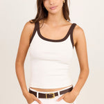 A woman modeling a white ribbed tank top with dark brown trim on the scoop neckline and armholes, paired with white pants and a brown belt, against a plain light beige studio background.