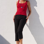 A woman wearing a bright red ribbed tank top, black cropped trousers, and black sandals leans against a white wall in harsh sunlight, casting a strong shadow.