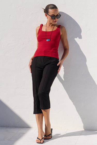 A woman wearing a bright red ribbed tank top, black cropped trousers, and black sandals leans against a white wall in harsh sunlight, casting a strong shadow.