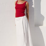 A woman wearing a red ribbed tank top and a long white skirt, standing against a white wall with strong sunlight and shadows.