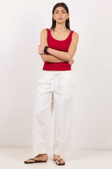 A woman stands against a plain white studio background wearing a sleeveless red tank top, loose white drawstring pants, and black flip-flop sandals, with her arms crossed.