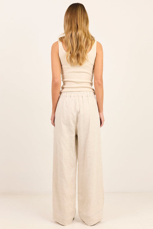 A woman with blonde hair is shown from the back, wearing a beige ribbed tank top and matching beige wide-leg linen pants against a plain off-white studio background.