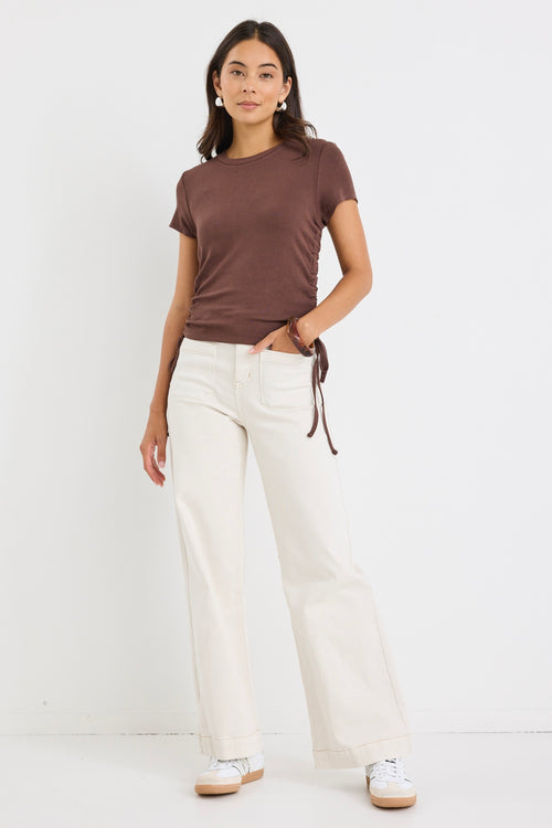 Woman wearing a fitted chocolate brown short sleeve tee with ribbed texture and adjustable side ruching, paired with high-rise cream jeans.