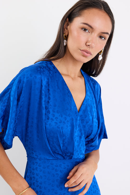 Woman wearing an electric blue satin jacquard midi dress with short kimono sleeves, a wrap-style bodice, and flowing skirt, standing against a white background