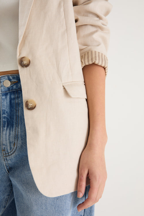 Woman wearing a long-sleeve cream linen blazer with button front and flap pockets, styled with a white top and light blue jeans.