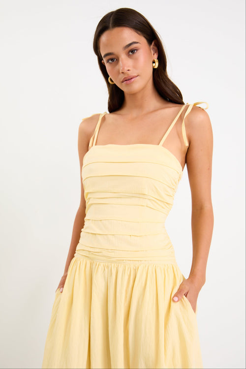 Model wearing a butter-yellow tie-strap maxi dress with a pleated bodice and flowing skirt