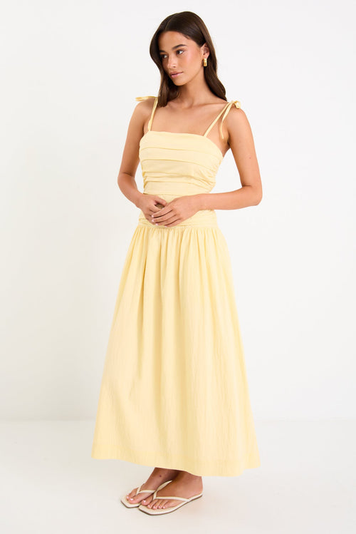 Model wearing a butter-yellow tie-strap maxi dress with a pleated bodice and flowing skirt