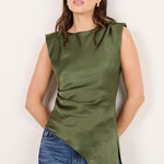 A woman modeling a sleeveless olive green asymmetrical top paired with blue jeans and black sandals, standing against a plain off-white studio background.
