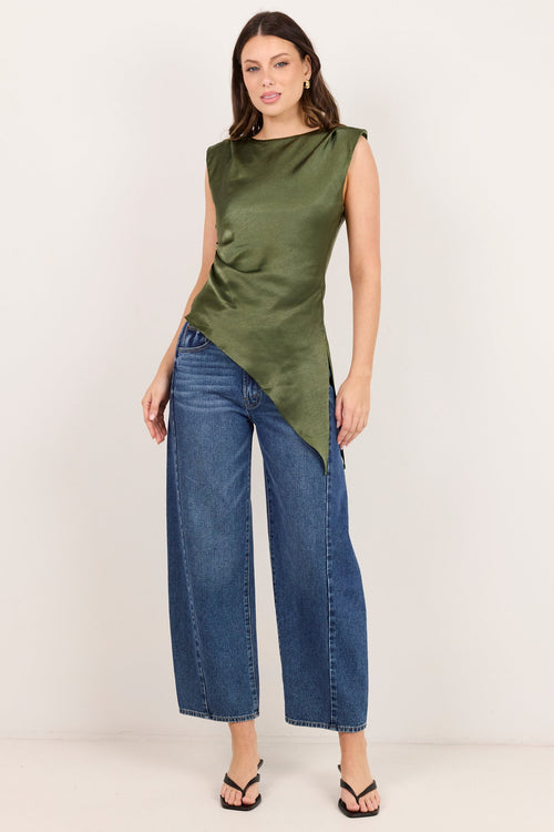 A woman modeling a sleeveless olive green asymmetrical top with blue wide-leg jeans and black sandals, standing against a plain off-white studio background.