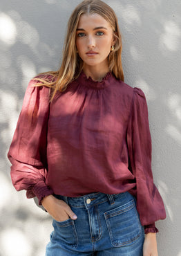 Woman wearing a burgundy blouse and blue jeans against a light gray wall.