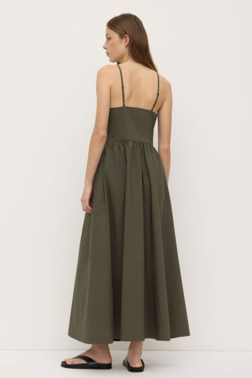 A woman wearing a sleeveless khaki green midi dress with a high neckline, fitted bodice, and gathered waist. The dress has side pockets and a relaxed A-line silhouette, styled simply with black sandals.’