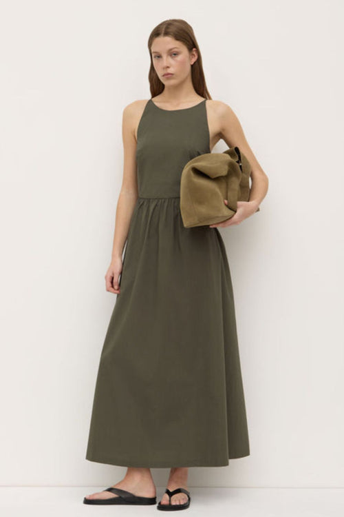 A woman wearing a sleeveless khaki green midi dress with a high neckline, fitted bodice, and gathered waist. The dress has side pockets and a relaxed A-line silhouette, styled simply with black sandals.’