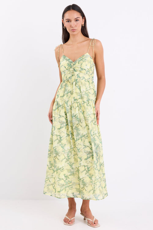 Woman wearing a yellow and green floral print maxi dress with adjustable tie straps, shirred back, and tiered skirt, standing against a white background.