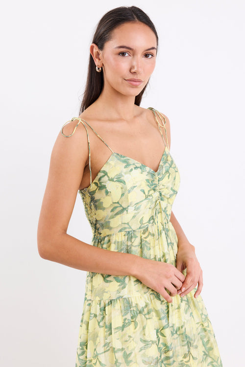 Woman wearing a yellow and green floral print maxi dress with adjustable tie straps, shirred back, and tiered skirt, standing against a white background.