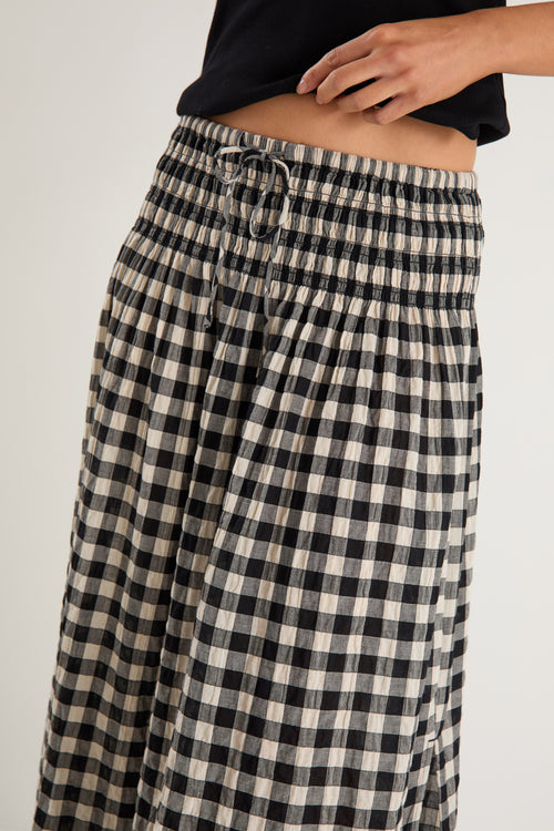Woman wearing a black gingham maxi skirt with shirred waist, tiered design, and side split, styled with light flat shoes.