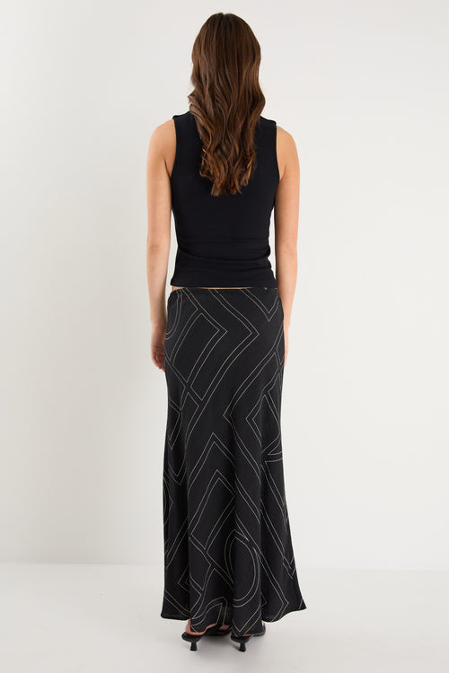 A model wears a long black skirt with an ivory geometric line print, featuring an elastic waist and a flowing silhouette, styled with a black sleeveless top and sandals.
