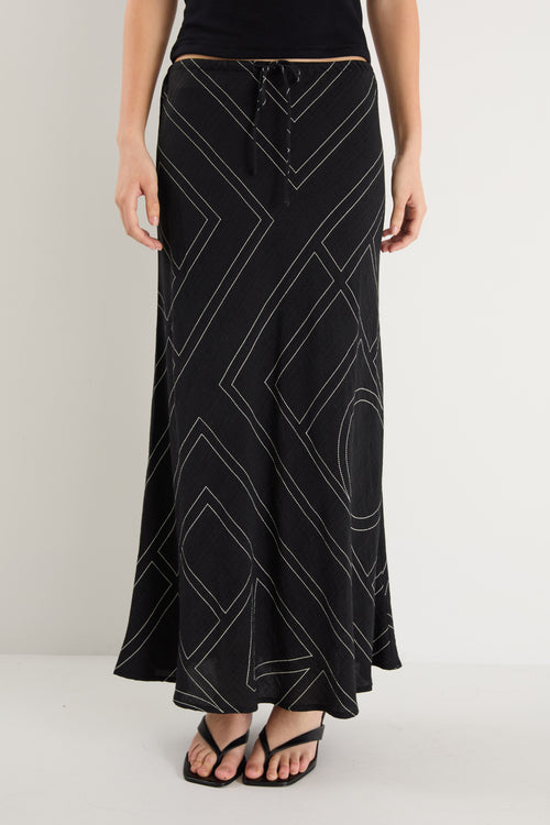 A model wears a long black skirt with an ivory geometric line print, featuring an elastic waist and a flowing silhouette, styled with a black sleeveless top and sandals.
col_pattern