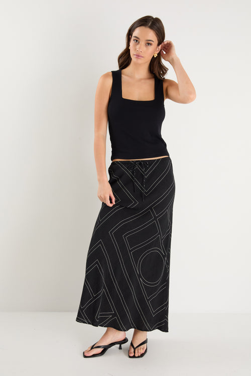 A model wears a long black skirt with an ivory geometric line print, featuring an elastic waist and a flowing silhouette, styled with a black sleeveless top and sandals.
