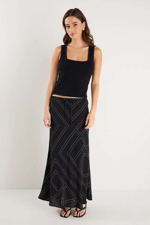 A model wears a long black skirt with an ivory geometric line print, featuring an elastic waist and a flowing silhouette, styled with a black sleeveless top and sandals.