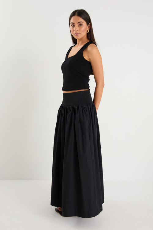 A model wears a black maxi skirt with a ribbed waistband and pleated design, styled with a black sleeveless top and sandals.
