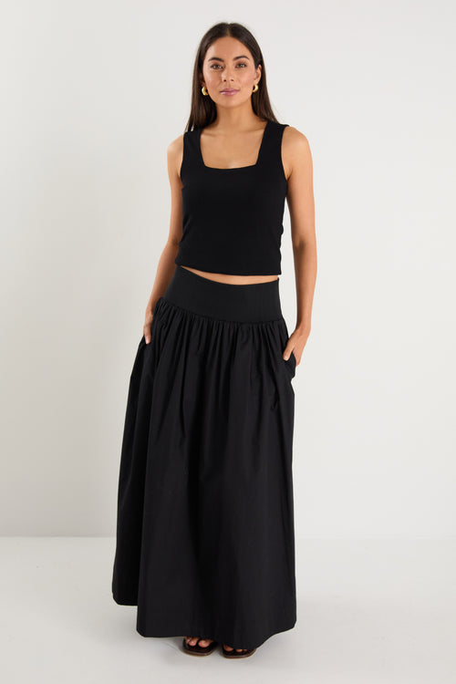 A model wears a black maxi skirt with a ribbed waistband and pleated design, styled with a black sleeveless top and sandals.