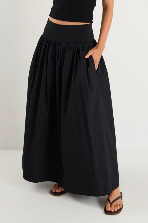 A model wears a black maxi skirt with a ribbed waistband and pleated design, styled with a black sleeveless top and sandals.